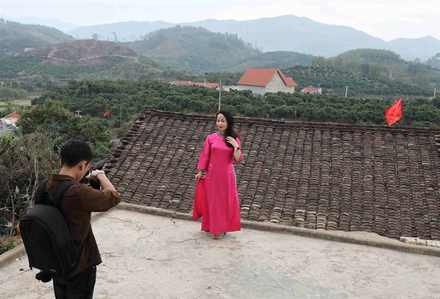 A tourist poses for a photo with the background featuring the yin-yang tiled roof of a Nùng traditional house. A tourist poses for a photo with the background featuring the yin-yang tiled roof of a Nùng traditional house.