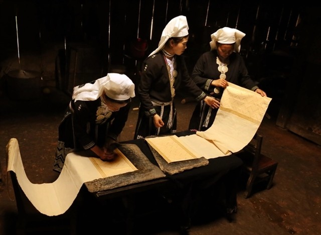 First, the white fabric is laid flat on a surface, such as a stone slab or table, and then smoothed and refined using a stone grinder. The pure beeswax, cut into small pieces for use, is placed in a bowl and heated over charcoal with a low flame to ensure the wax remains hot enough to adhere to the fabric and create clear patterns.  Depending on the predetermined patterns for the hem of the skirt, collar or sleeves, the women use various tools to dip into the beeswax and print the designs onto the fabric.